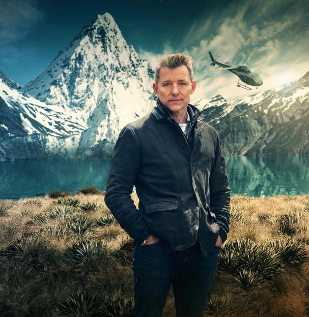 Ben Shephard standing in front of a lake with snowy mountains and a helicopter in the background.