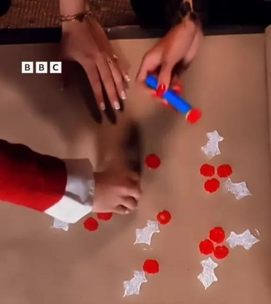 BBC logo on plain brown wrapping paper, with two sets of hands making holly patterns with red paint.