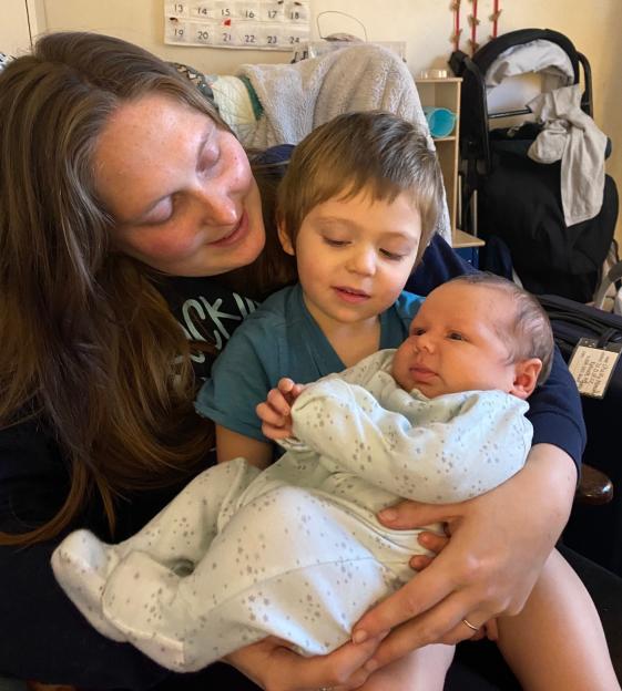 A smiling woman with long brown hair holds a baby in a white, star-patterned onesie while a young boy wearing a blue shirt leans in to look at the baby.
