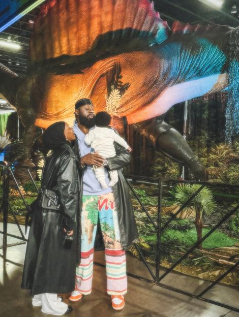 A woman in a hijab and long black coat stands beside a man holding a baby, in front of a dinosaur display.