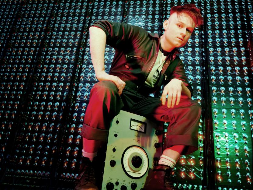 LOOK MUM NO COMPUTER, an artist with red hair, sits on a vintage sound mixing board with a background of glowing circuit boards.