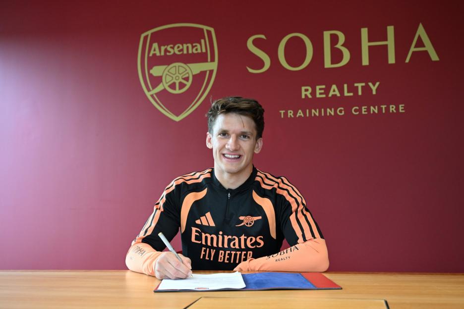 Christos Tzolis signing paperwork at a desk, wearing an Arsenal training top, with the Arsenal logo and "SOBHA REALTY TRAINING CENTRE" on the red wall behind him.