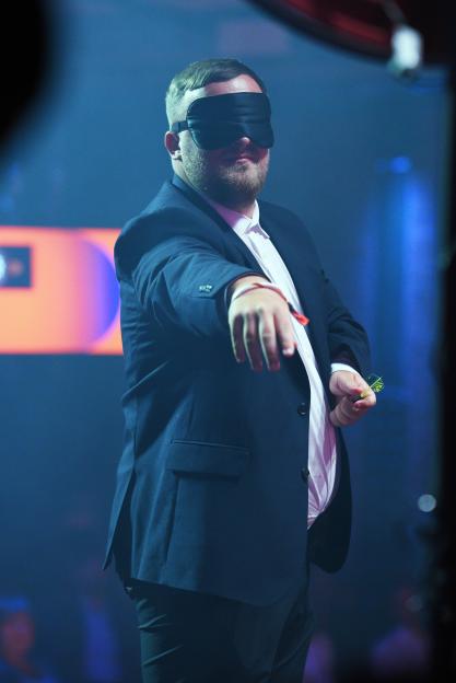 Darts player Luke Littler blindfolded with a sleep mask, extending his arm and holding a dart.