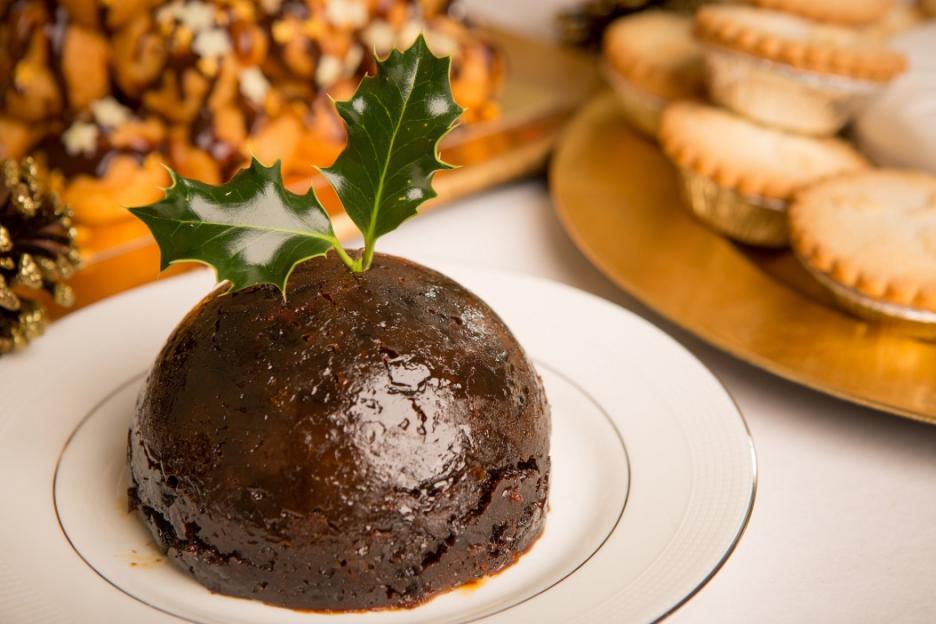 Christmas pudding with holly and a gold pinecone, with mince pies in the background.