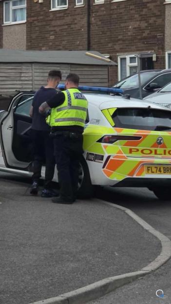 A police officer in a high-visibility vest stands next to a man by an open police car door, with residential buildings in the background.