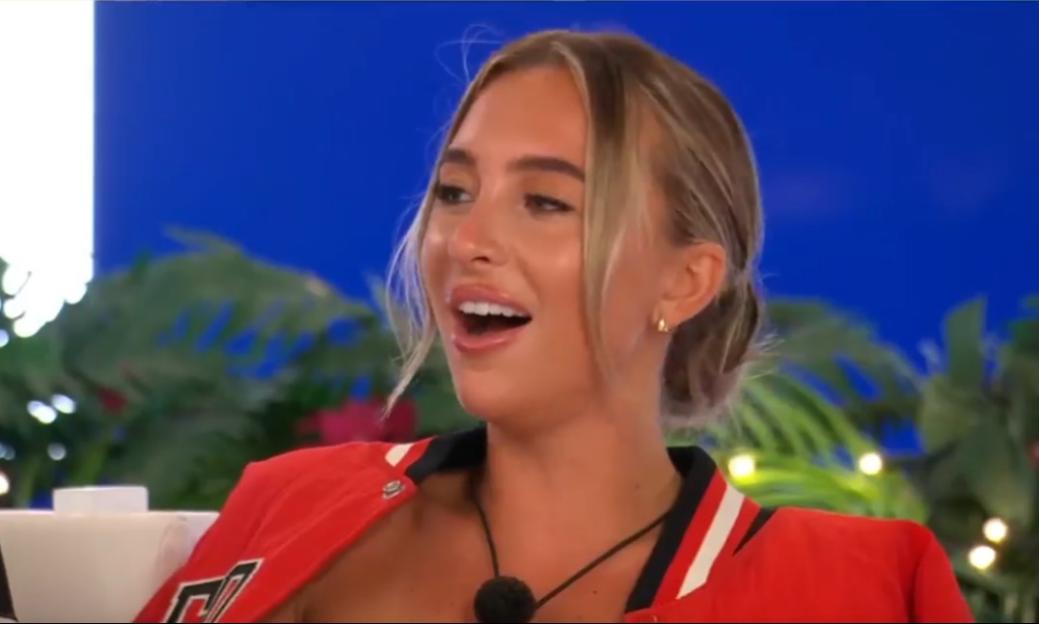 A woman with blonde hair in a red jacket, looking surprised with her mouth open.