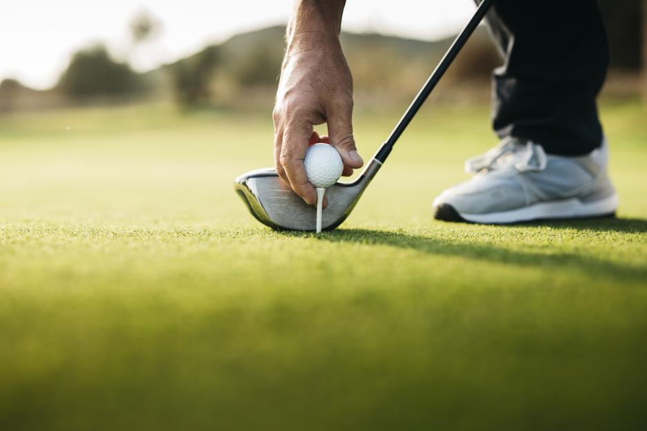 A golfer's gloved hand places a golf ball on a tee with a driver resting beside it.