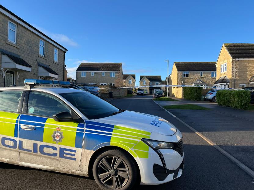 A West Yorkshire Police car parked on a residential street with police tape visible in the background.