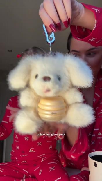 A person holding up a white fluffy dog keychain with a honey pot in its stomach, with another person in red pajamas in the background.