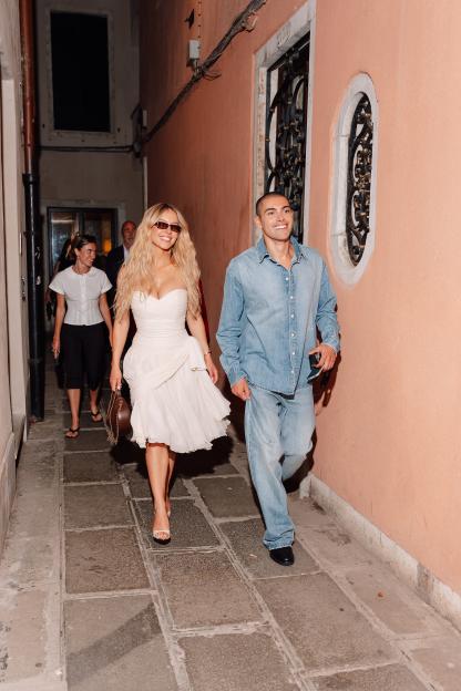 Sydney Sweeney in a white dress and black sunglasses, smiling and walking next to a man in a denim shirt and jeans.