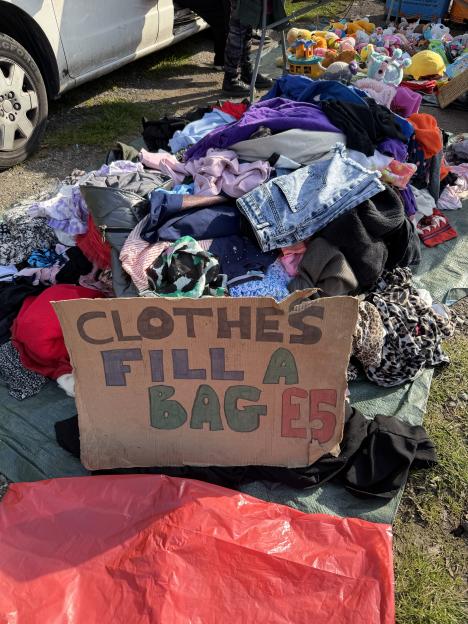 A car boot sale stall with a large pile of clothes and a cardboard sign reading "Clothes Fill A Bag £5."