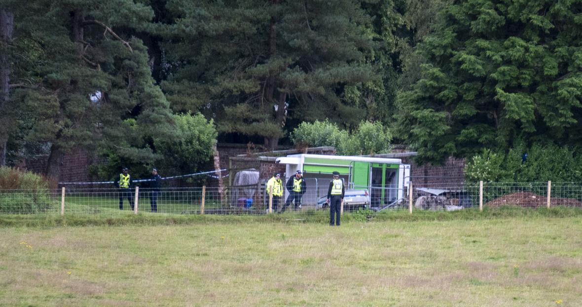 Police officers are seen at a rural property, with a green trailer and wooded area in the background, possibly investigating a suspected murder-suicide.