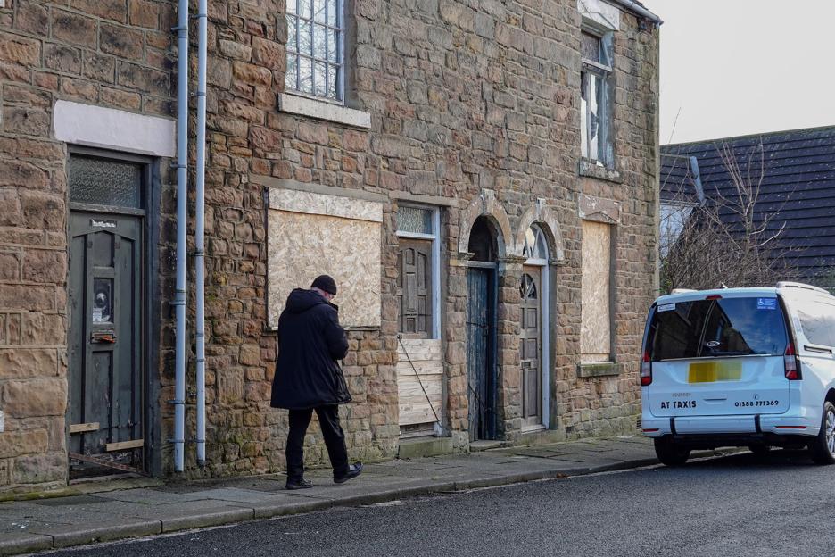 A person walking past boarded-up stone buildings next to an "AT TAXIS" van.