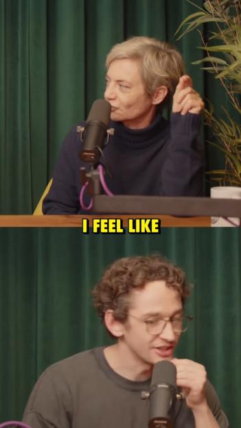 Two podcast hosts sitting at a table with microphones, the top one is a woman with short blonde hair in a navy blue sweater, and the bottom one is a man with curly brown hair wearing glasses.