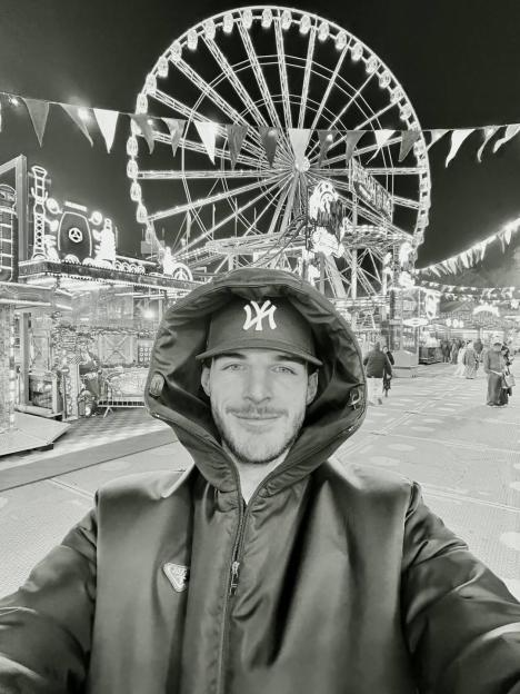 A man smiling in a hooded jacket and baseball cap at an amusement park with a Ferris wheel in the background.
