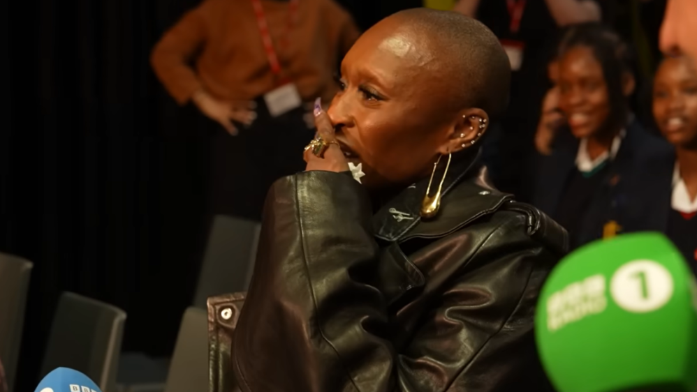 A person with a shaved head and a black leather jacket crying with their hand to their face.