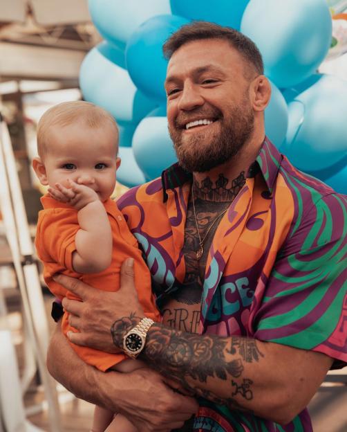 Conor McGregor holding his baby, Rian, who is wearing an orange shirt and looking at the camera.