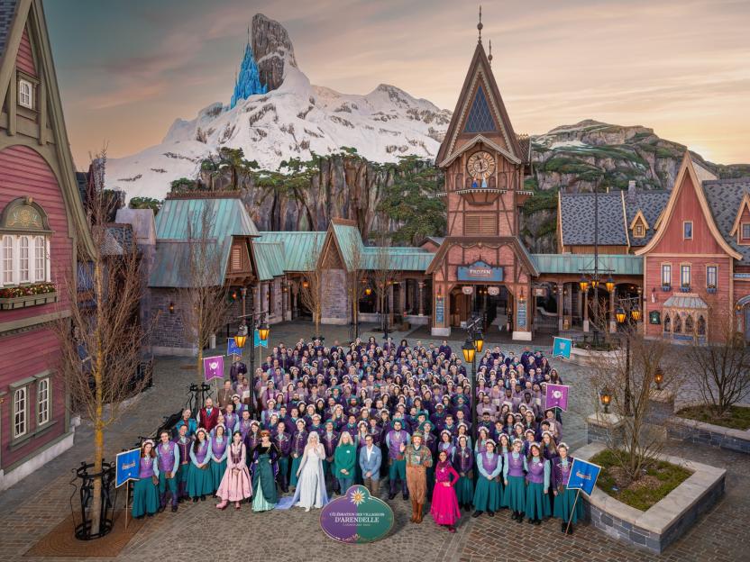 A large group of people dressed in costumes from the film Frozen stand in front of buildings designed to look like they are from Arendelle, with a snow-covered mountain in the background.
