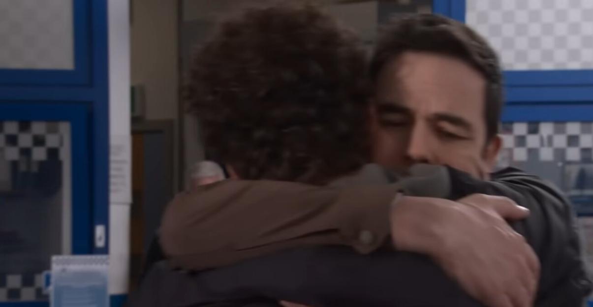 Two men embrace, one with his back to the camera and the other with his face visible.