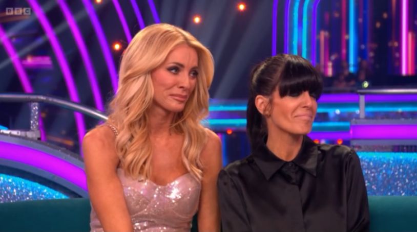 Two women, one in a sequined dress and the other in a black top, sitting on a blue couch against a backdrop of purple and blue stage lights.