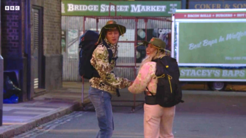 Two people wearing hiking gear and hats holding hands on a street.