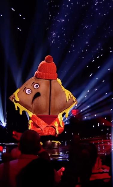 The Masked Singer character Toastie, a grilled cheese sandwich wearing a red beanie, stands on stage with spotlights.