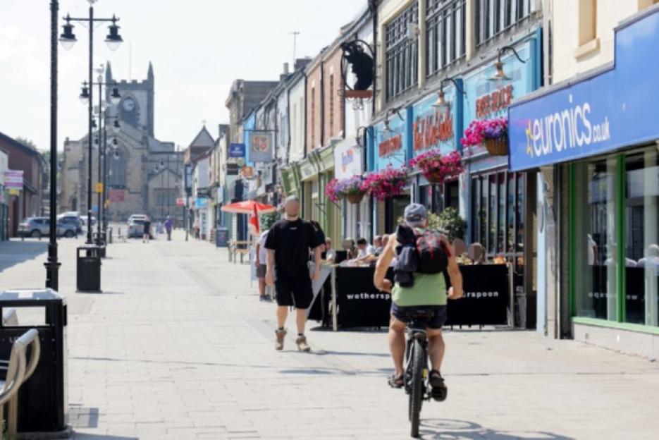 A street in Seaham, County Durham, with stores and a church tower visible in the background, as people walk along a paved sidewalk and sit at an outdoor cafe.