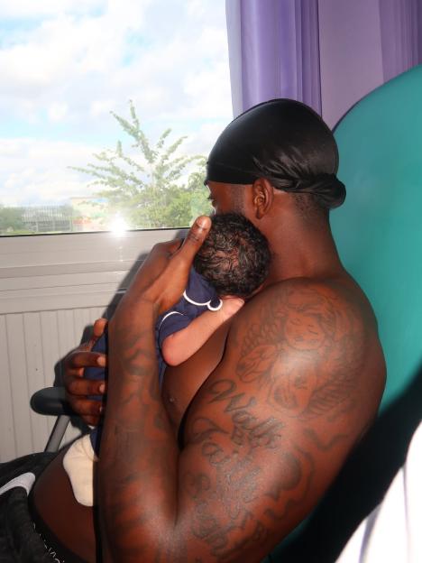 A person with a durag and tattoos on their arm holds a baby on their chest, looking out a window.