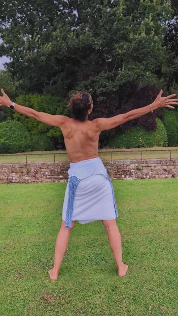 A person with their back to the camera, arms outstretched, wearing a skirt and no top, standing barefoot on grass.