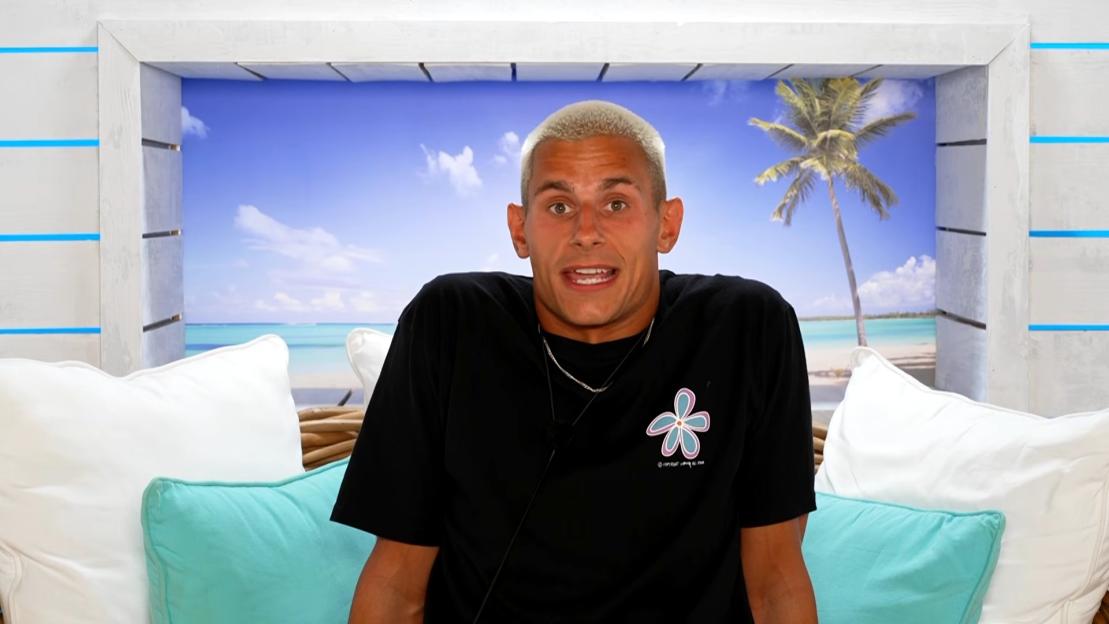 A man with blond hair and a black t-shirt with a blue and purple flower design looks surprised while sitting on a couch.
