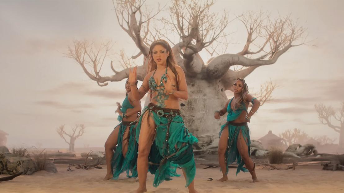 Shakira performing with two backup dancers in a desert landscape with bare trees.