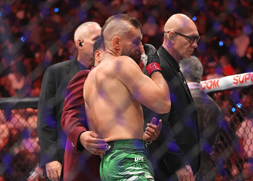 A bare-chested fighter with a shaved head and green shorts wipes his face with a gloved hand while being escorted by officials.