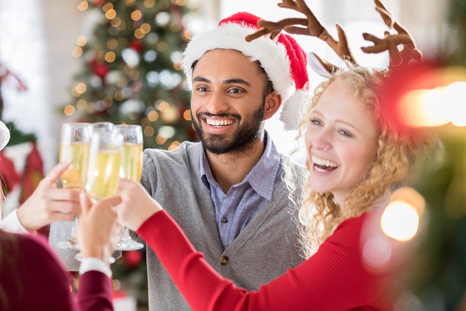 Man in Santa hat and woman in reindeer antlers clinking champagne flutes with friends.