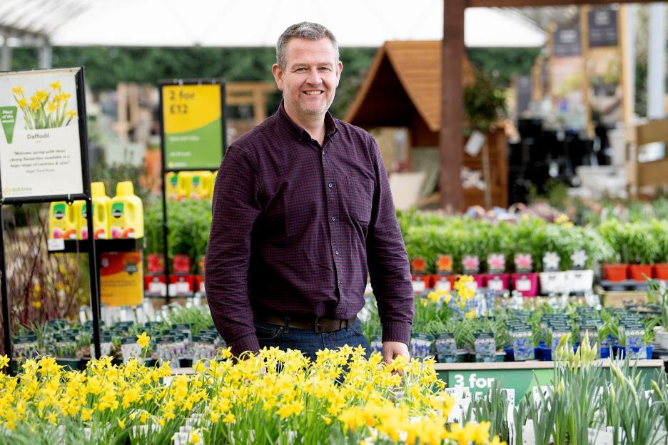 A man in a checkered shirt smiling in a Dobbies Garden Centre surrounded by daffodil plants.