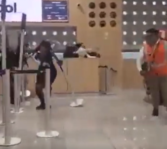 A person in a dark outfit with what appears to be a staff or weapon, and a person in an orange vest, in what appears to be an airport or large venue.