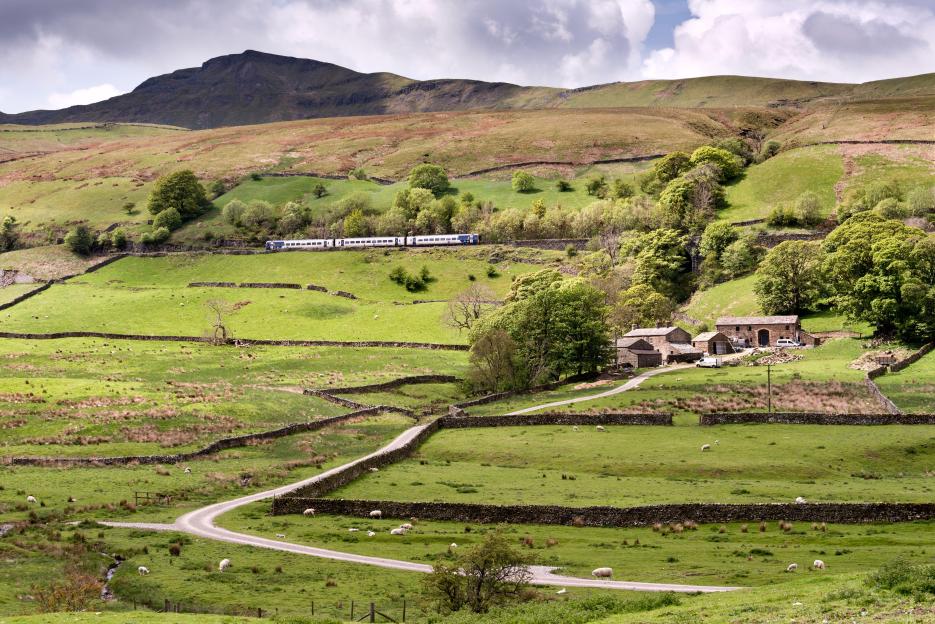 A passenger train travels along the Settle-Carlisle railway below Wild Boar Fell and above the hamlet of Outhgill, Mallerstang, Westmorland, UK.