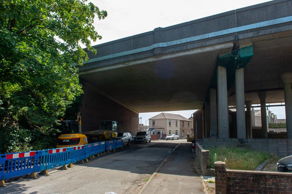 A street view underneath the M4 motorway bridge in Port Talbot, showing two large yellow construction vehicles behind blue and white safety barriers.