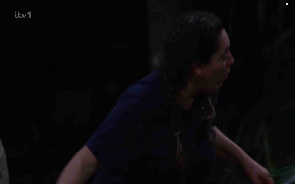 A person wearing a dark blue shirt and long hair is shown in a dark room.