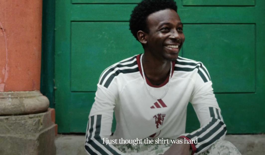A smiling Black man in a white Adidas long-sleeved shirt with black stripes on the shoulders and a Manchester United logo.