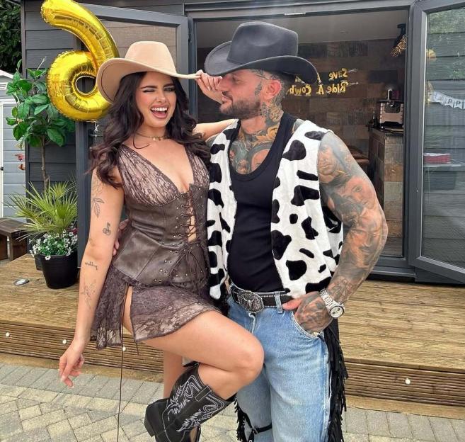 A woman in a brown dress and cowboy boots, and a tattooed man in a cow-print vest and jeans with a cowboy hat.