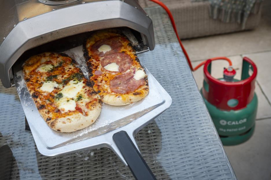 Two pizzas on a peel, one pesto and one pepperoni, coming out of a pizza oven powered by a Calor gas tank.