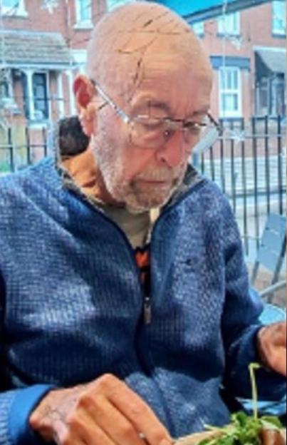 A man with a cracked skull effect on his head and a beard, wearing glasses and a blue zip-up top, is looking down at a plate of food.