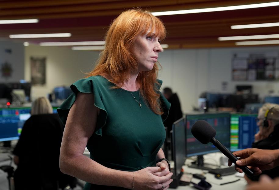A woman with red hair and a green dress speaks into a microphone in an office setting.