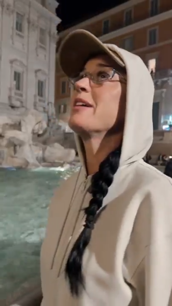 Katy Perry in a hoodie and baseball cap, looking up at the Trevi Fountain in Rome.