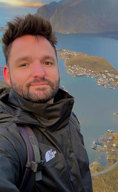 Man in a black jacket and backpack taking a selfie with a mountain and village on a lake in the background.