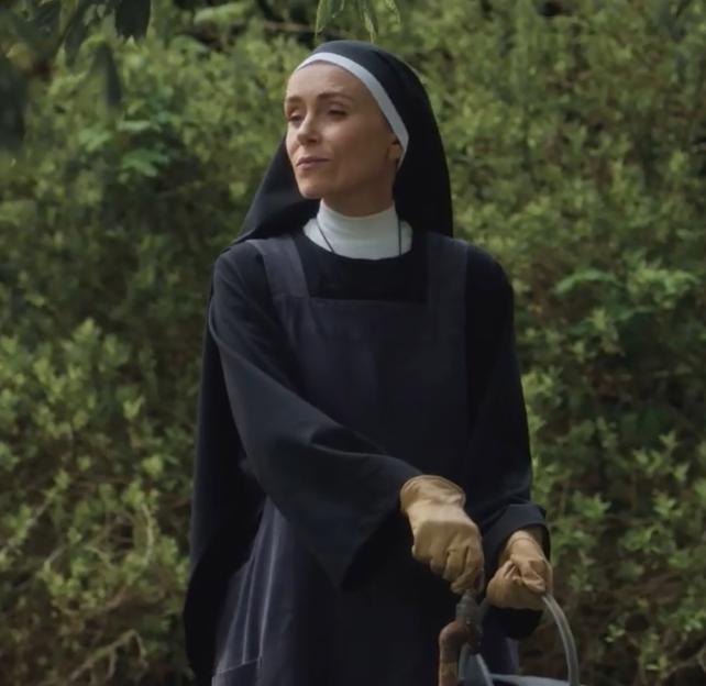 A nun in a black habit and apron, wearing gardening gloves, holds a watering can handle, looking towards the left with a slight smile.