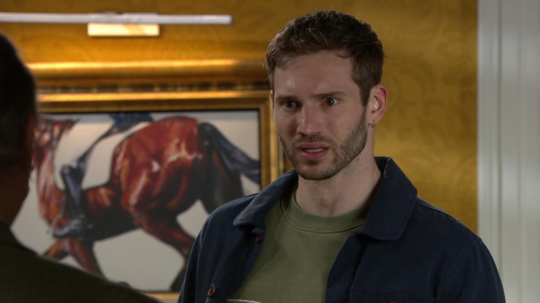 A man with a beard and dark hair wearing a dark blue jacket and green shirt looks wide-eyed, with a painting of a racehorse in the background.