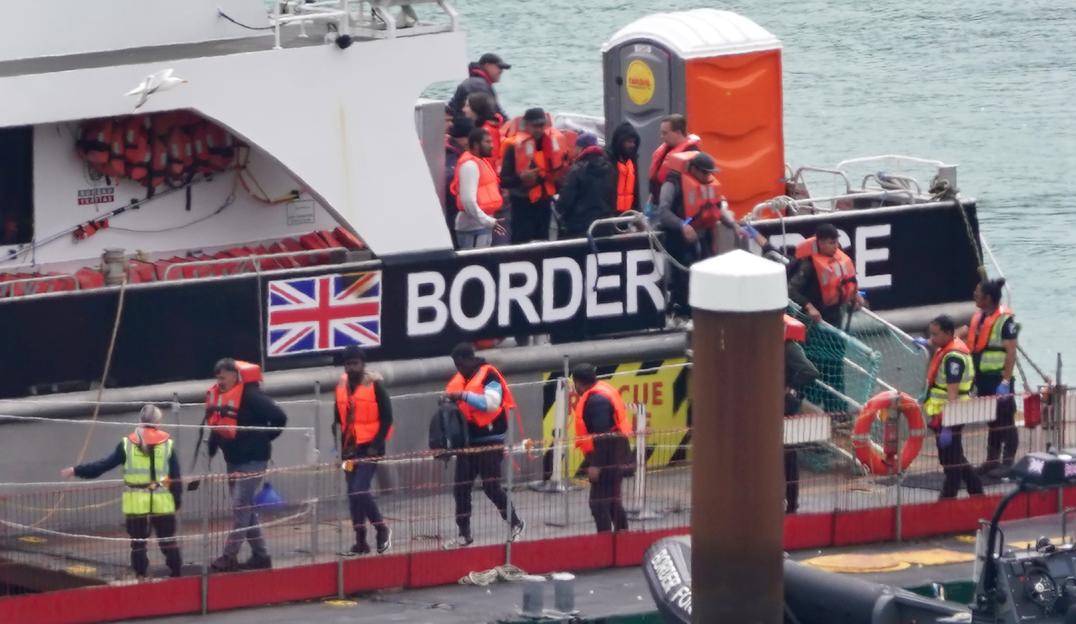 A group of people wearing orange life vests are disembarking from a Border Force vessel with a Union Jack flag on its side.
