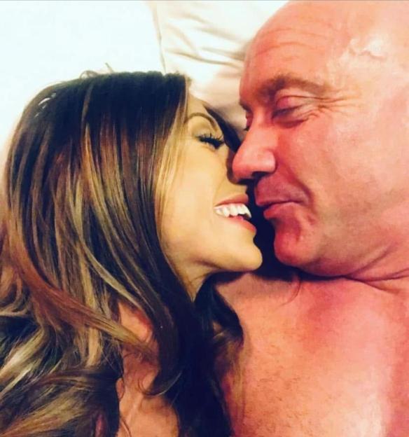 A young woman with long, brown hair and an older, bald man lying in bed, nose to nose, with the woman smiling broadly.