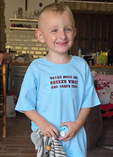Dax Hammill, age five, smiling while wearing a light blue t-shirt that says "VACAY MODE ON: BREEZY VIBES AND SANDY TIDES".